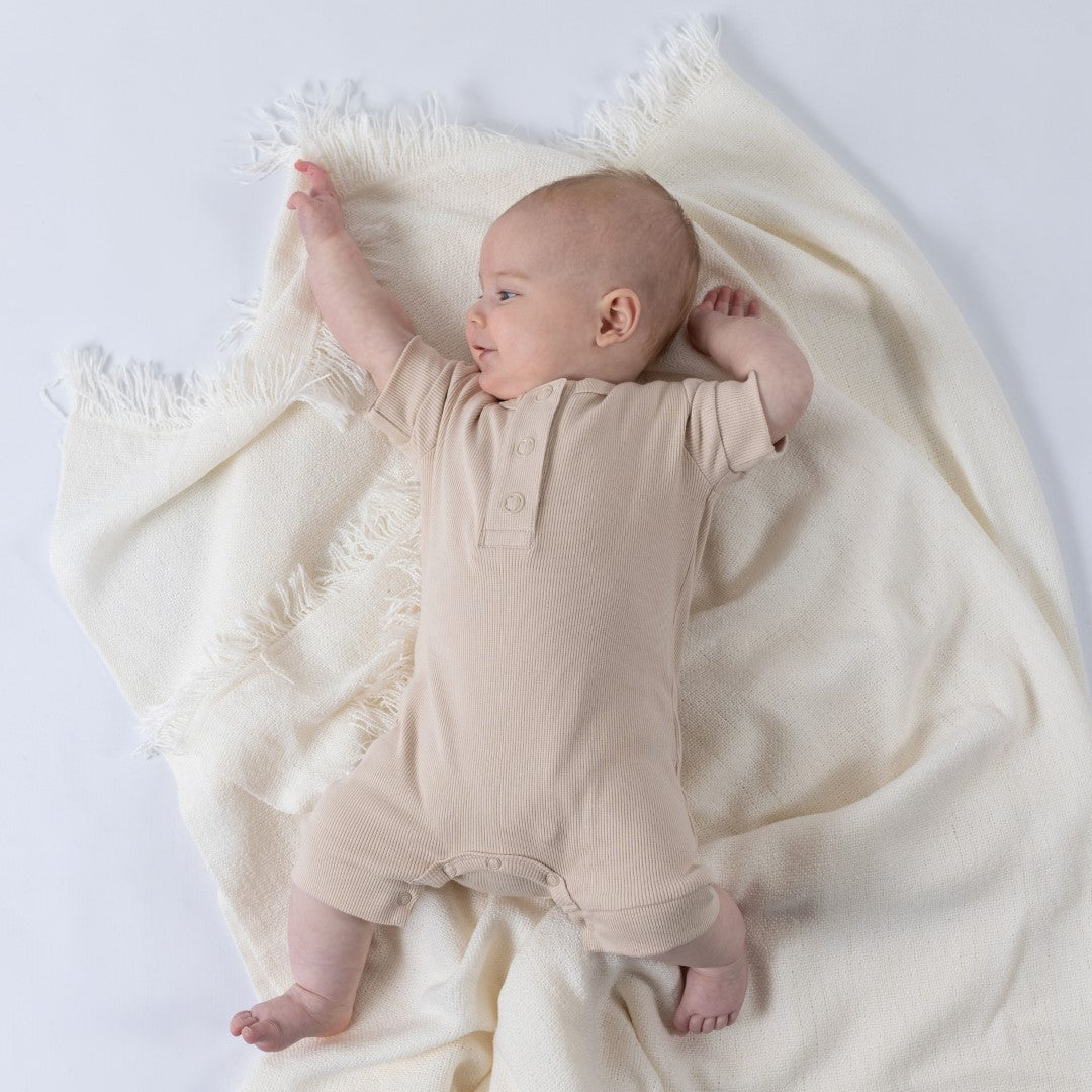 Baby laying on a cream blanket, wearing an oat short sleeve rib baby romper, looking to the side.