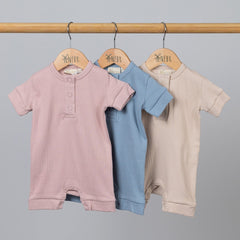 Three short sleeve rib baby rompers hanging on a wooden frame in dusty pink, slate blue, and oat colours.
