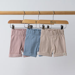 Three pairs of ribbed organic cotton kids shorts in oat, dusty pink, and slate blue hanging on wooden hangers, set against a neutral background.