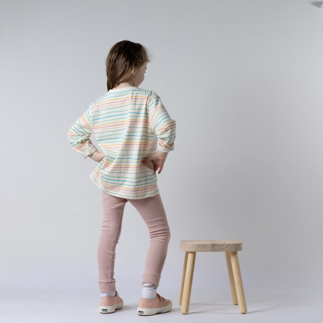 Rear view of a girl standing beside a wooden stool, wearing a kids long sleeve striped t-shirt in soft pastel colours and dusty pink leggings.