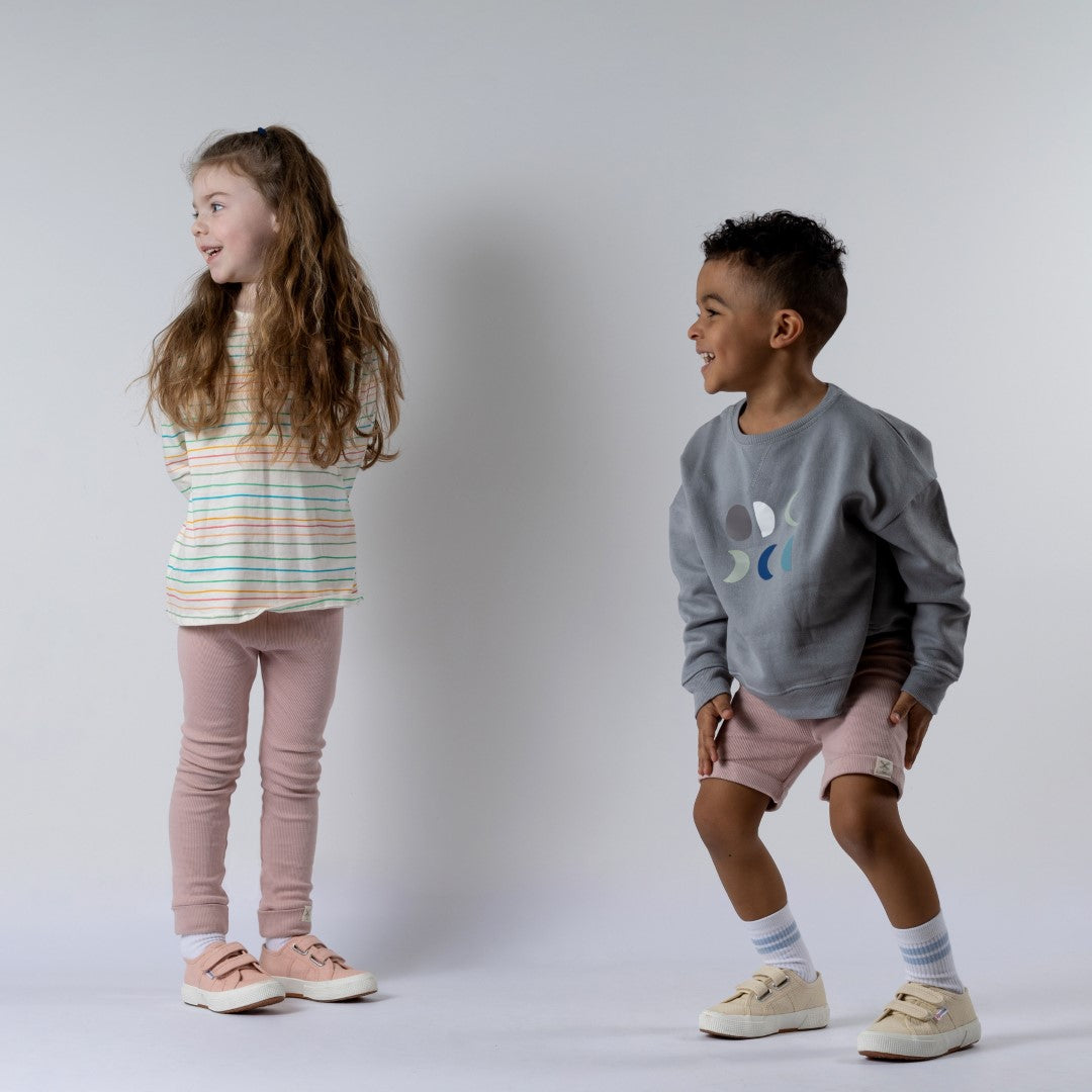 Girl wearing a kids long sleeve striped t-shirt in pastel colours and dusty pink leggings stands beside a boy in a moon-print jumper and pink shorts.